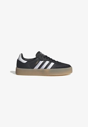 Zapatilla deportiva de cuero negro con tres rayas plateadas, punta texturizada y suela de goma. Cuenta con un collar acolchado y un diseño con cordones.