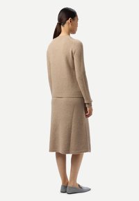 Kvinna med mörkt hår uppsatt, klädd i en beige stickad tröja och matchande knälång kjol, står med ryggen mot betraktaren och bär gråa flatskor.