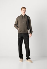 Felpa verde oliva con polsini a coste, dettaglio del logo sul petto, abbinata a jeans scuri e sneaker beige, completata da una piccola borsa a tracolla.