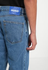 Jeans de mezclilla azul claro con un bolsillo trasero que incluye una etiqueta azul con la palabra "HUGO". Los jeans tienen un corte relajado y un ligero desgaste.