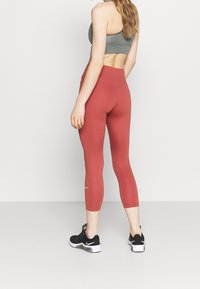 Leggings taille haute, ajustés, dans une teinte corail avec une texture lisse, arborant un petit logo blanc sur la jambe inférieure. Associés à des baskets noires.