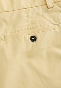 Gros plan sur un tissu beige avec une poche arrière fermée par un bouton noir et un détail de couture renforcée.