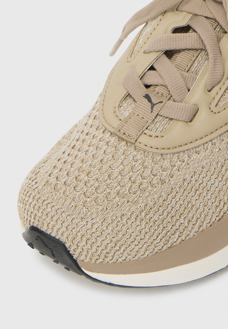 Zapatilla deportiva beige con parte superior de malla texturizada, cierre de cordones y lengüeta acolchada; cuenta con un acento negro cerca de la suela.