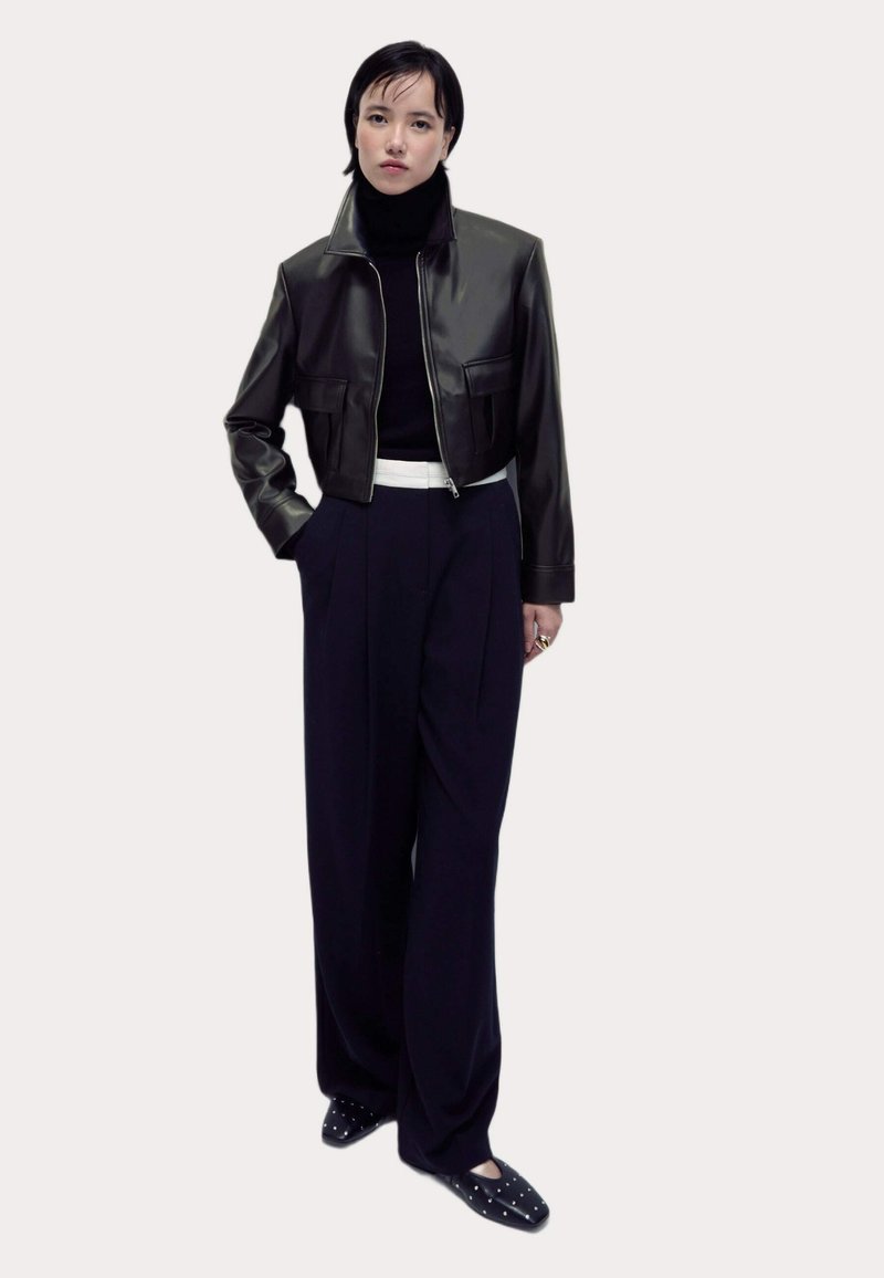 Schwarze Leder-Cropped-Jacke mit hohem Kragen, kombiniert mit dunkelblauen, hochgeschnittenen Hosen und schwarzen Ballerinas mit silbernen Nieten.