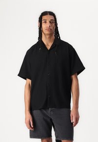 Camisa negra de manga corta con botones y textura acanalada, con un cuello con muescas y cinco botones negros. Combinada con pantalones cortos grises.