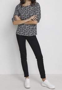 Modella in piedi con le braccia incrociate, indossa un top a fantasia bianca e nera, pantaloni neri e sneakers bianche su uno sfondo neutro.