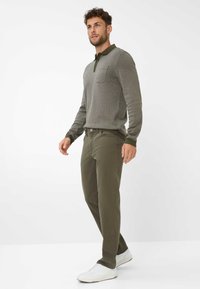 Pantaloni verde oliva con gamba dritta, abbinati a una polo grigia a maniche lunghe con colletto scuro e taschino sul petto. Sneakers bianche.