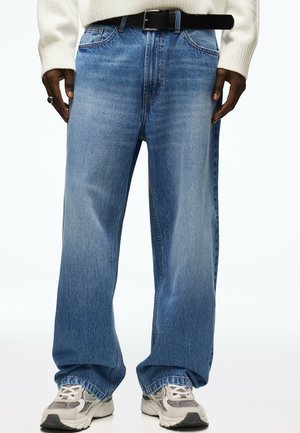 Person iført løse blå jeans, hvide sneakers, beige sweater, sort bælte og en ring på venstre hånd mod en hvid baggrund.