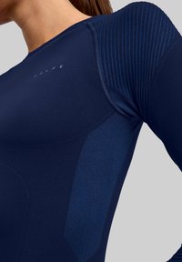 Granatowa sportowa bluzka z długim rękawem z teksturowanymi panelami i prążkowanymi akcentami. Charakteryzuje się ergonomicznym krojem oraz kontrastującymi matowymi i błyszczącymi wykończeniami.