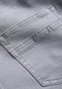 Šedá deninová látka s detailní kapsou s kontrastním šitím a hladkým povrchem, zdůrazňující upravený, strukturovaný design.