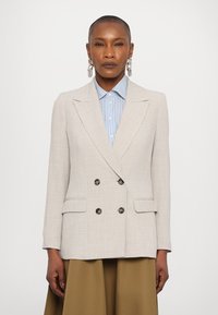 Lys beige dobbeltspent blazer laget av teksturert stoff, med store svarte knapper og normal krage, kombinert med en lyseblå stripete skjorte.