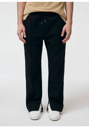 Osoba ubrana w czarne szerokie spodnie z przeszyciami z przodu i białe trampki, stojąca na jednolitym białym tle.