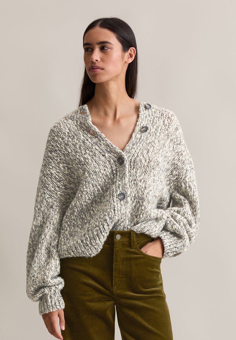 Cárdigan de punto grueso en gris y blanco con escote en V, grandes botones grises y puños acanalados, combinado con pantalones de pana verde.