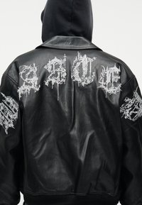 Persona con chaqueta de cuero negra con letras góticas blancas intrincadas bordadas en la espalda y las mangas, con la capucha negra puesta.