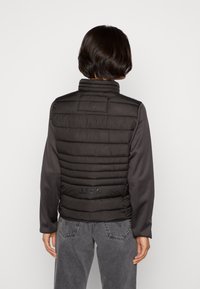 Gilet a vento nero con design trapuntato, colletto alto e logo in tonalità sul retro, indossato sopra una maglietta grigia a maniche lunghe e jeans.