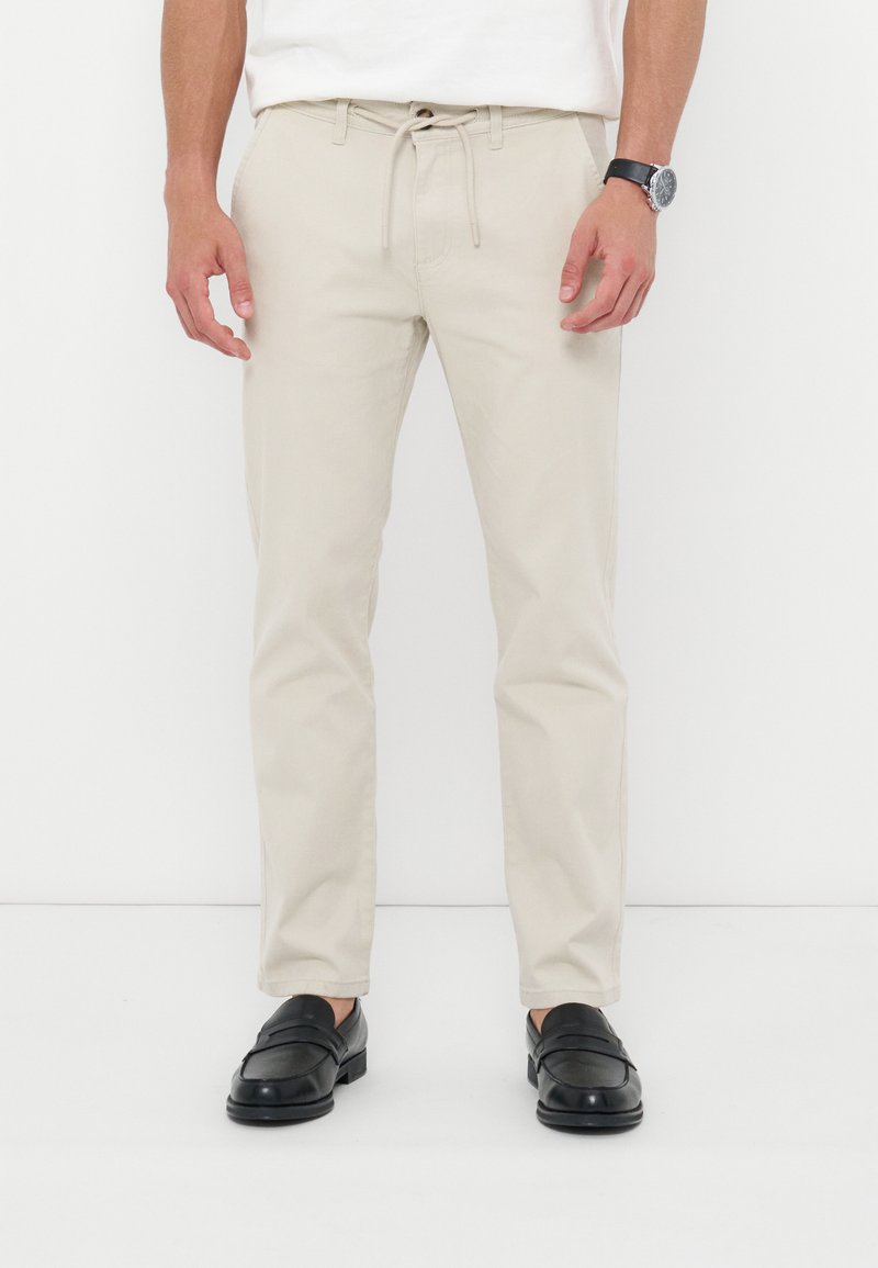 Homme portant un pantalon beige à cordon, des mocassins noirs, une chemise blanche et une montre noire au poignet, debout devant un fond blanc.