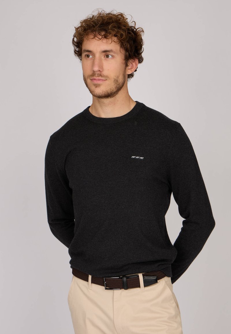 Pull gris foncé à manches longues avec un col rond et une finition texturée, arborant un petit logo blanc sur la poitrine, associé à un pantalon beige clair et une ceinture marron.