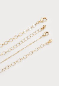 Złote metalowe łańcuchy o różnorodnym wzornictwie: jeden z heksagonalnymi ogniwami, drugi z okrągłymi ogniwami, a trzeci z zapięciem typu homar. Gładka tekstura.