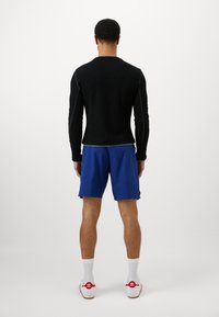 Top de manga longa preto e calções azuis, com um design justo. Meias brancas e sapatilhas com detalhes em vermelho. Fundo branco liso.