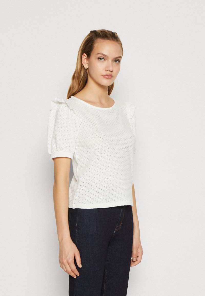 Top blanco de manga corta con un patrón texturizado y acentos con volantes en los hombros, combinado con jeans de mezclilla oscura.