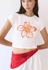 Witte cropped t-shirt met een print van een oranje hibiscusbloem; gecombineerd met een doorschijnende rode wikkel om de taille. Soepele, lichte stof.