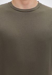Close-up of a person wearing an olive green crew neck t-shirt on a white background.