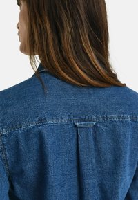 Chemise en denim de couleur bleue, avec un col pointu, un détail de plis au dos et des coutures visibles. Le matériau est doux avec une légère texture.