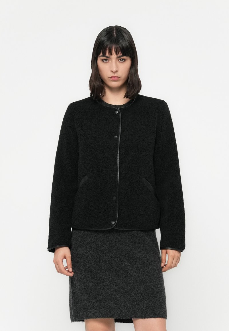 Femme aux cheveux noirs raides portant une veste en polaire noire à boutons et une jupe gris foncé, se tenant contre un fond blanc.