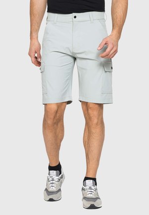Uomo che indossa pantaloncini cargo grigio chiaro con tasche laterali con bottoni, calzini neri alla caviglia, scarpe da ginnastica grigie e una maglietta nera su uno sfondo uniforme.
