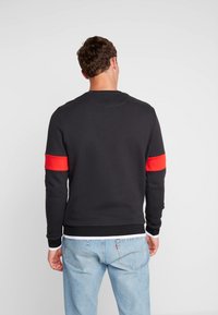 Felpa nera con strisce rosse accentate sulle maniche, polsini e fascia in vita a coste, realizzata in un tessuto liscio, stile aderente, vista posteriore.