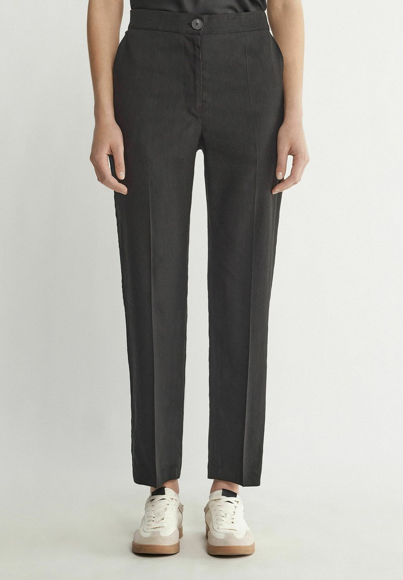 Personne portant un pantalon noir cintré et des baskets blanches, debout sur un fond clair uni, les mains détendues le long du corps.