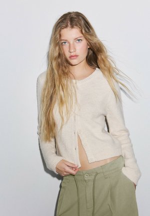 Jeune femme aux longs cheveux blonds portant un pull crème boutonné et un pantalon vert olive, debout devant un fond blanc uni.