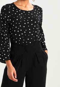 Blusa nera con pois bianchi, scollatura rotonda e lunghe maniche a balze. Abbinata a pantaloni neri a vita alta con pieghe frontali.