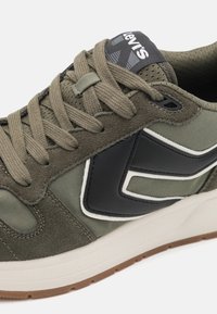 Zapatillas en verde oliva con materiales de ante y lienzo, que presentan un logo negro texturizado, detalles en blanco y una suela de goma.