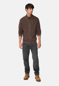 Braune gestrickte Zip-up-Strickjacke mit Rippbündchen und Saum, kombiniert mit dunklen Jeans und hellbraunen Arbeitsstiefeln. Einfaches, strukturiertes Design.