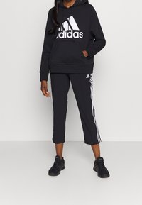 Černá adidas mikina s velkým bílým logem, doplněná o černé zkrácené kalhoty s bílými bočními pruhy. Nosené s černými sportovními teniskami.