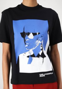Melnā īsā piedurkņu T-krekls ar zilu un balto grafisko portretu, kas valkā saulesbrilles. Apakšā teksts "KARL LAGERFELD JEANS".