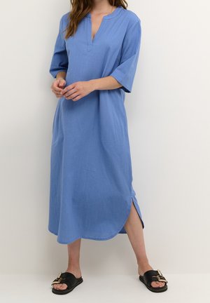 Femme portant une robe bleue ample mi-longue avec des manches au niveau des coudes et une fente latérale, associée à des sandales noires à enfiler avec des boucles dorées.