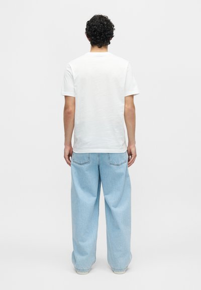 Άνδρας με σγουρά μαλλιά που φοράει απλό λευκό μπλουζάκι και φαρδιά ανοιχτό μπλε τζιν, στεκόμενος με την πλάτη προς ένα απλό φόντο.