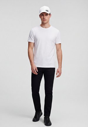 Nuori mies seisoo päällään valkoinen t-paita, mustat housut, mustat tennarit ja valkoinen lippis, jossa on musta auton kuvake.