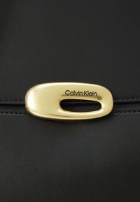Cuir noir avec un tirette de fermeture éclair ovale dorée arborant le logo "Calvin Klein" en texte noir, rehaussé de détails cousus.