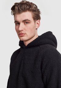 Felpa con cappuccio nera in pile con una superficie texturizzata e morbida, dotata di un ampio cappuccio e una vestibilità rilassata, progettata per comfort e calore.