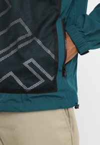 Tyrkysová lehká bunda s černou síťovanou panelou a geometrickými bílými vzory, elastickými manžetami a boční kapsou na zip.