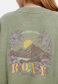 Berglandschap met zon, omringd door paarse bloemen, en tekst "ROXY Sinds 1990" op olijfgroen stof.