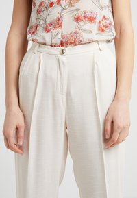 Jasno kremowe spodnie w plisy z teksturowanej tkaniny. Posiadają zapięcie na guzik z przodu oraz boczne kieszenie, zaprezentowane z kwiecistą bluzką.