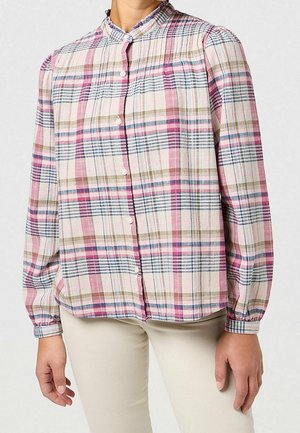 Kobieta nosząca długorękawową, zapinaną na guziki bluzkę w różowo-fioletowo-zielono-niebieską kratę, w połączeniu z jasnobeżowymi spodniami.