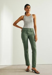 Zelené cargo kalhoty se slim fit střihem, vybavené předními kapsami a knoflíkovým zapínáním, kombinované s šedým tílkem a černými sandály na podpatku.