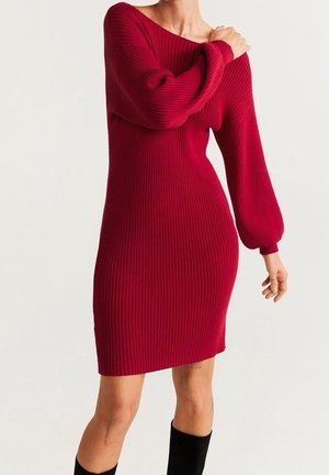 Femme portant une robe rouge moulante en maille côtelée à manches longues bouffantes, associée à des bottes noires montantes jusqu'au genou.