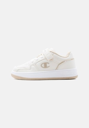 White low-top sneaker with beige accents, a strap closure, perforated details, and a logo on the side and sole.
