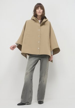 Beige Kapuzenumhang mit Knopfleiste, zwei großen Taschen und weiten Ärmeln, kombiniert mit ausgewaschenen grauen Schlagjeans und schwarzem Schuhwerk.